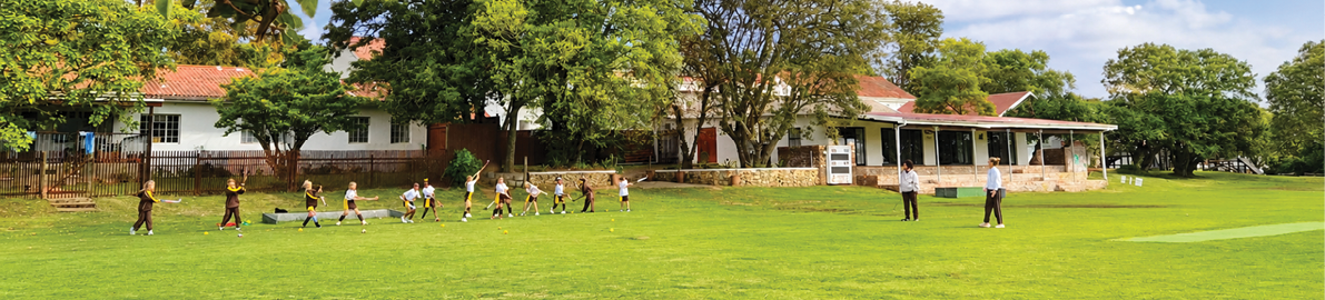 A group of children walking in a line across a grassy field with two adults nearby and houses and trees in the background. Description generated by AI