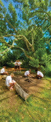 Five people in white shirts and shorts are gathered outdoors near a campfire setup under a large tree with green foliage on a sunny day. Description generated by AI