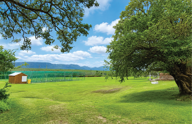 A large grassy field with a small wooden shed, a swing hanging from a tree, and a backdrop of mountains under a partly cloudy sky. Description generated by AI