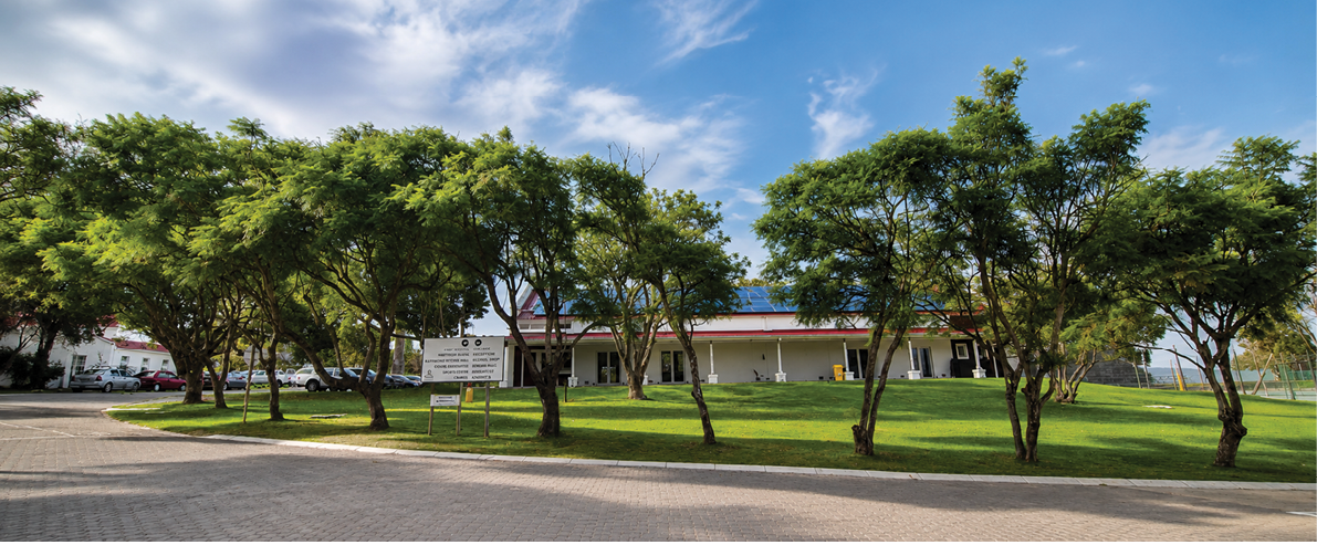A single-story building with a red roof is surrounded by green grass and several trees under a partly cloudy blue sky. Description generated by AI